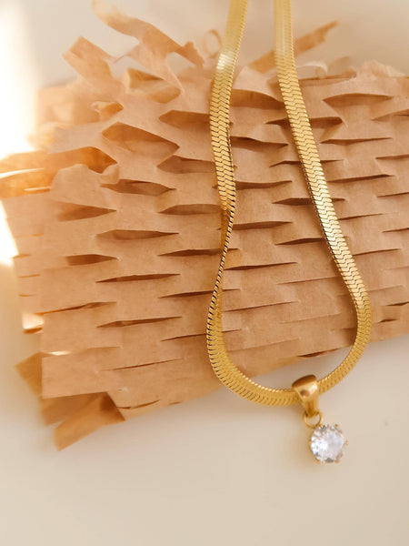 Flat lay: Close-up of 18K gold snake chain necklace with cubic zirconia round pendant on white surface