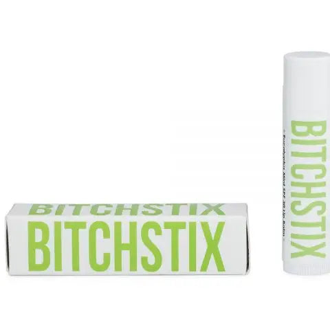 A picture showing two views of a BITCHSTIX lip balm product, with the packaging and the balm itself displayed. The packaging is primarily white with green text and accents, and the balm is white with green text and accents as well.
